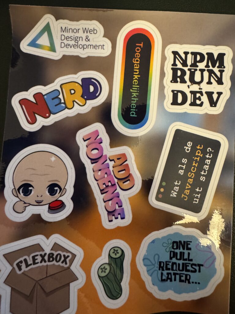 A glossy sheet of developer-themed stickers arranged on a dark surface. The stickers include: “Minor Web Design & Development” with a triangular logo; a vertical rainbow sticker reading “Accessibility” (translated from the Dutch “Toegankelijkheid”); “NPM RUN DEV” in bold lettering; a colorful “NERD” sticker; a bald cartoon character pressing a red button; a sticker reading “ADD NONSENSE”; a terminal-style sticker saying “What if JavaScript is turned off?” (translated from Dutch); a cardboard box labeled “Flexbox”; a small green pickle illustration; and a blue cloud-shaped sticker inspired by Spongebob that says “One pull request later…”. The overall theme is web development and programming culture.